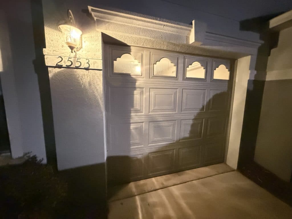 A suburban garage with a partially open new garage door in the foreground, showcasing modern design and sleek materials. A middle-aged contractor in professional work attire examines the door, holding a measuring tape. In the middle ground, tools like a drill and a ladder are neatly arranged outside the garage, hinting at recent installation work. The background features a well-kept driveway and a suburban neighborhood with green trees and houses under a bright blue sky, suggesting a pleasant day. Natural sunlight illuminates the scene, creating a warm and inviting atmosphere. The image captures the essence of home improvement and the importance of quality garage doors. Garaqge Door Replacement - How Much To Replace Garage Door?